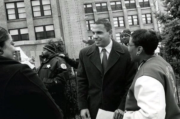 Ron Huberman standing outdoors in a coat and tie, smiling while speaking with people in front of a building