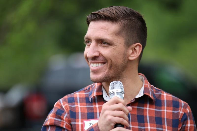 Zak Ringelstein speaking into a microphone outdoors, wearing a plaid shirt with a blurred green background.